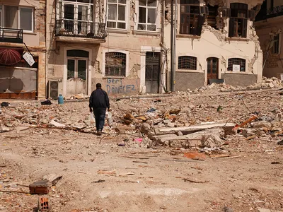 From behind, a person walks through a street filled with gray rubble, toward run-down buildings with graffiti in Turkish on the walls.