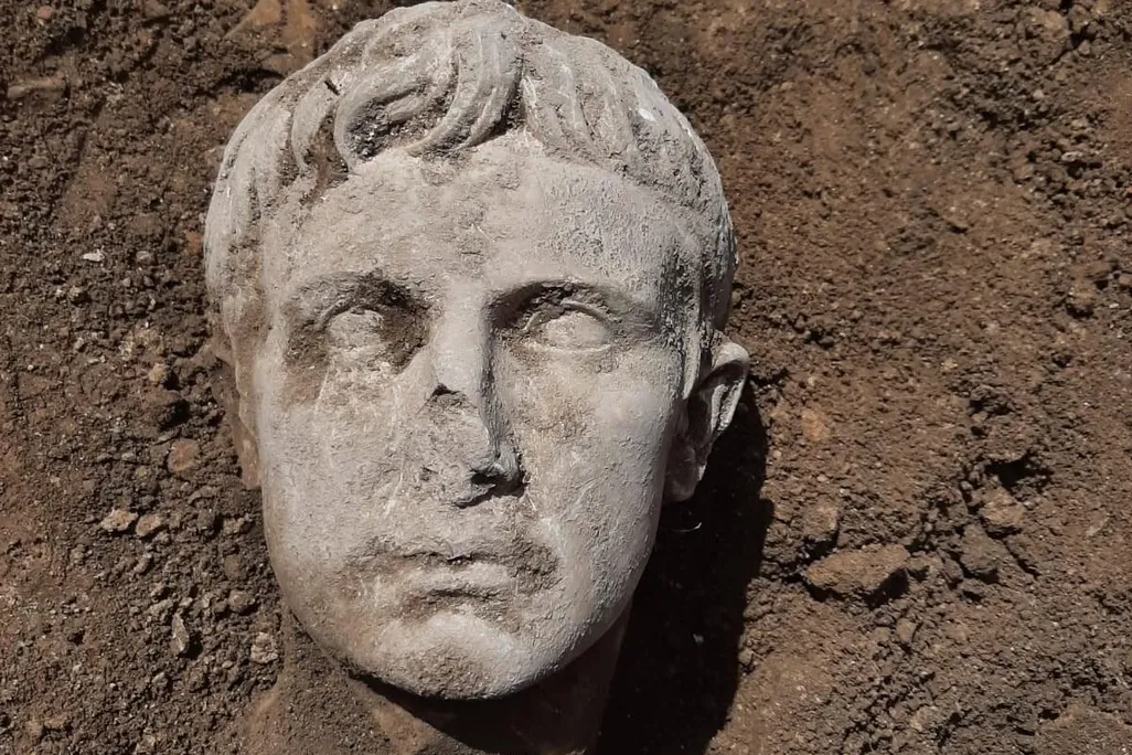 A white marble head covered partially in dirt; rests on a dirt ground next to someone's shoe