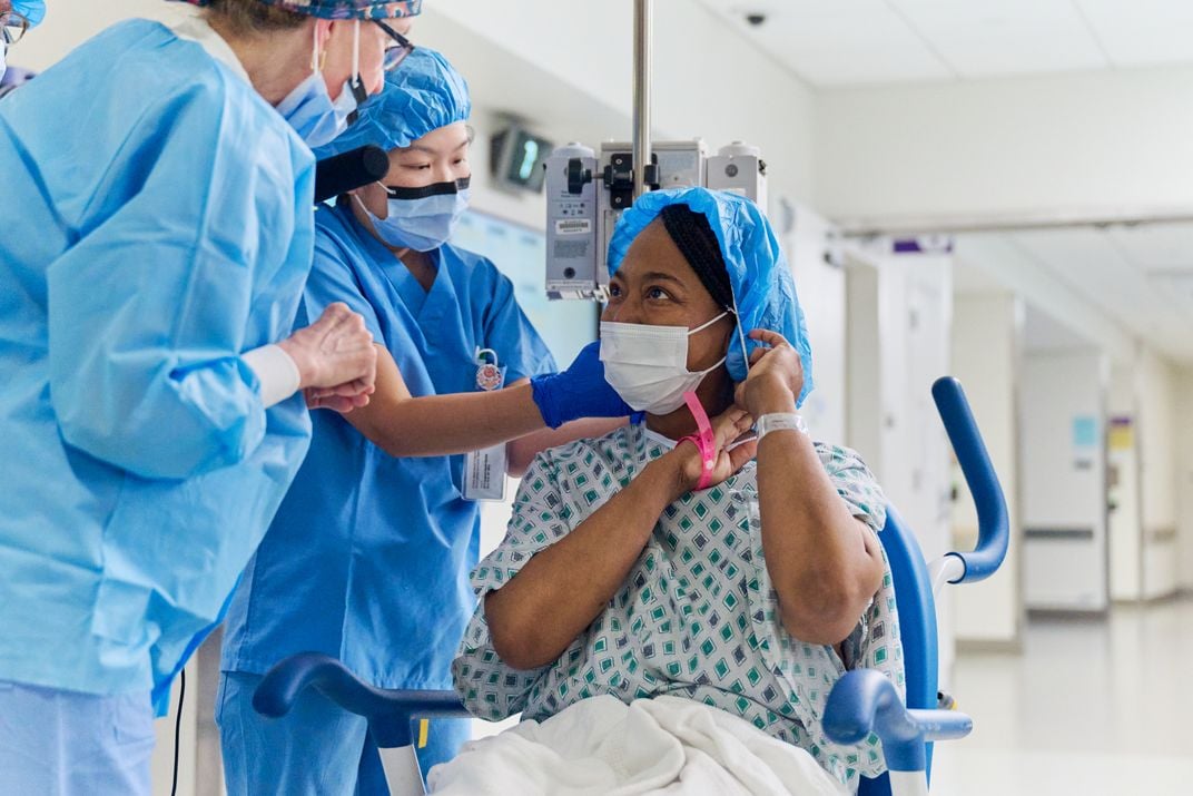 Woman wearing hospital gown and cap sitting in chair next to people wearing blue scrubs