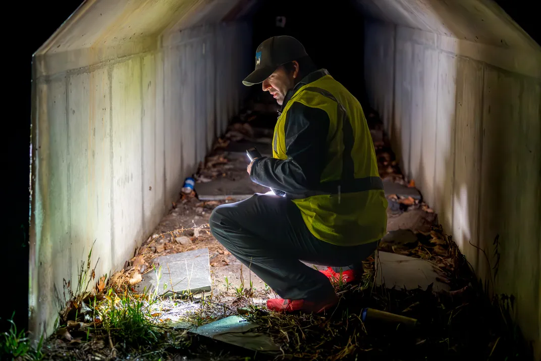 a man in a reflective vest squats with a light in an amphibian tunnel at night