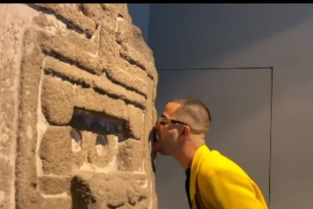 In a screenshot, a man in a yellow jacket licks the carved stone facade of an ancient work of Indigenous art