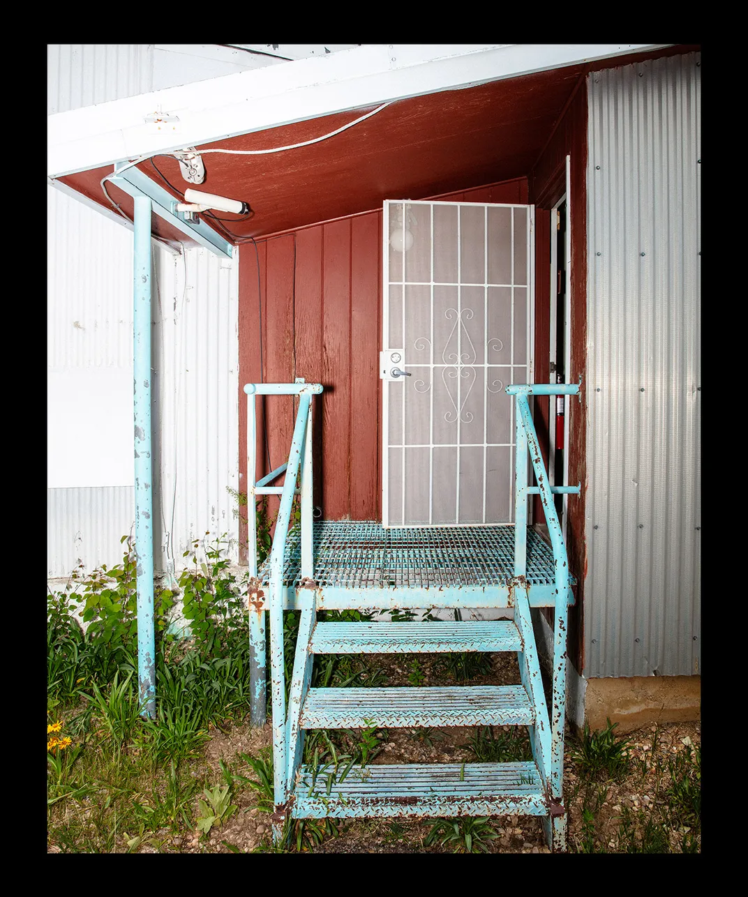 a blue metal staircase with peeling paint leads into a red-painted building with a white metal door