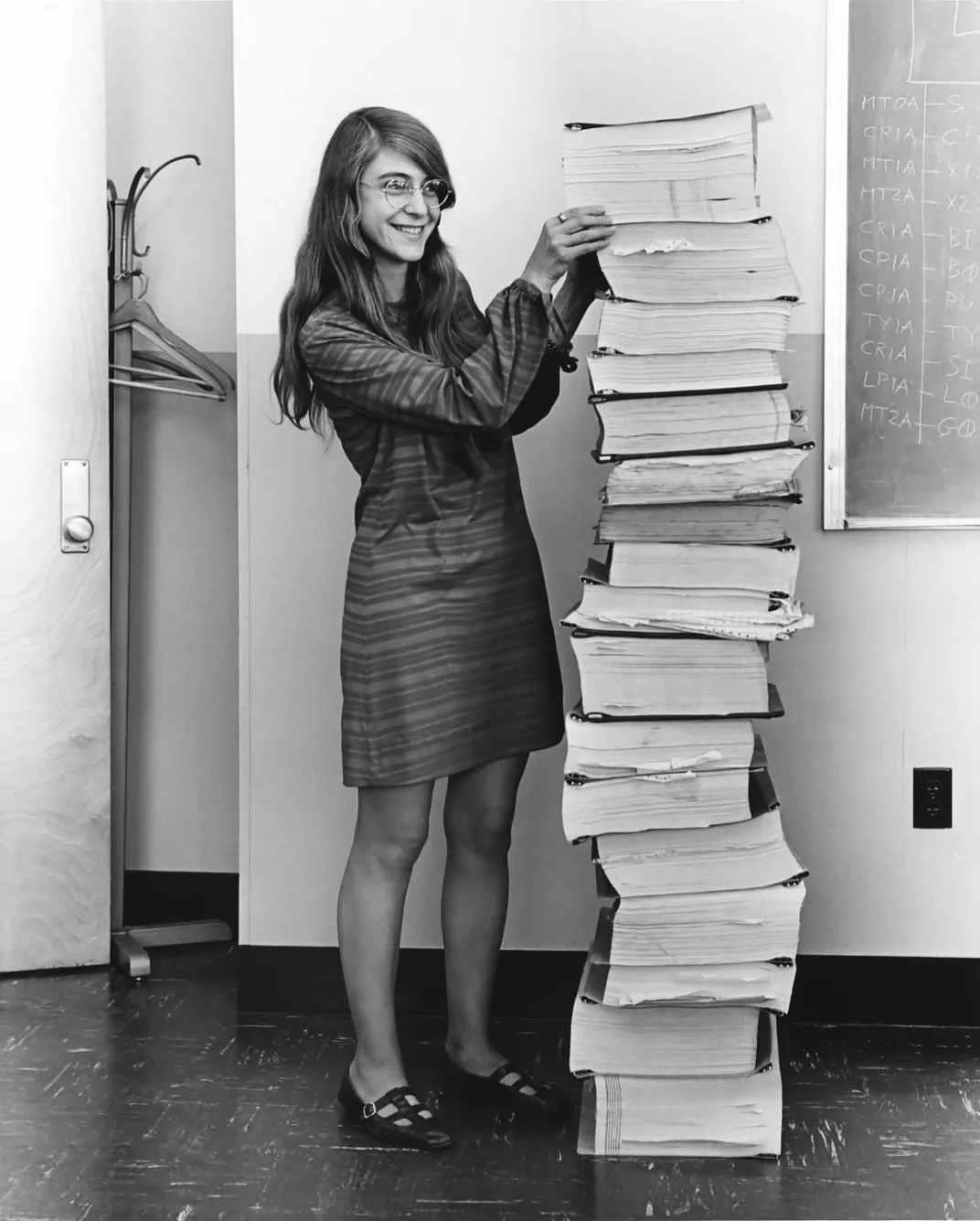 A black-and-white image of a long-haired women next to a giant pile of paper almost as tall as she is