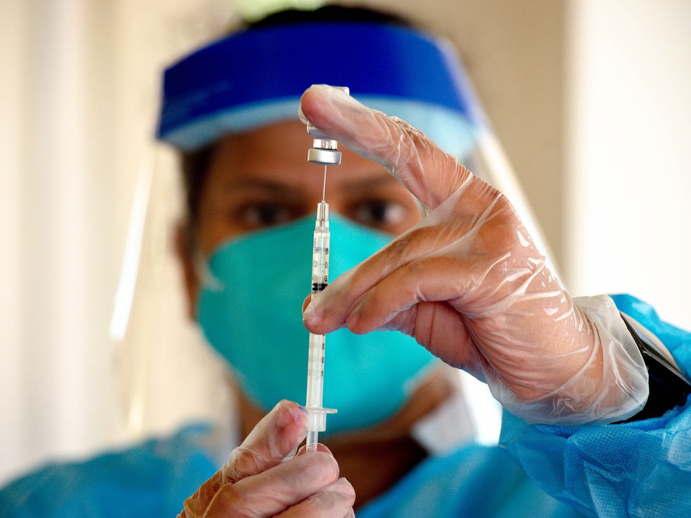 A pharmacist holds a vial of vaccine and a syringe in front of their face