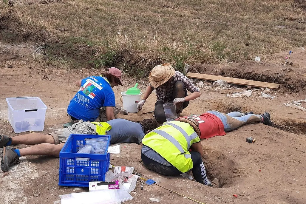 A group of four people clad in hats and reflective gear kneel and lie on their stomachs, peering into a hole dug into the dirt