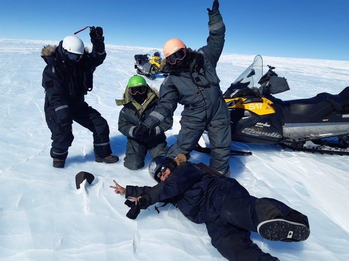 two researchers in winter coats stand with arms raised, one crouches behind the space rock, one lies on ground making peace sign