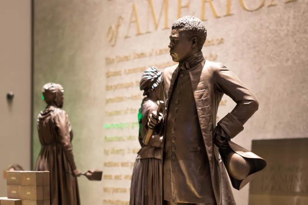 A bronze colored statue of Bannecker, a Black man wearing a suit jacket and high boots, stands in a museum exhibition
