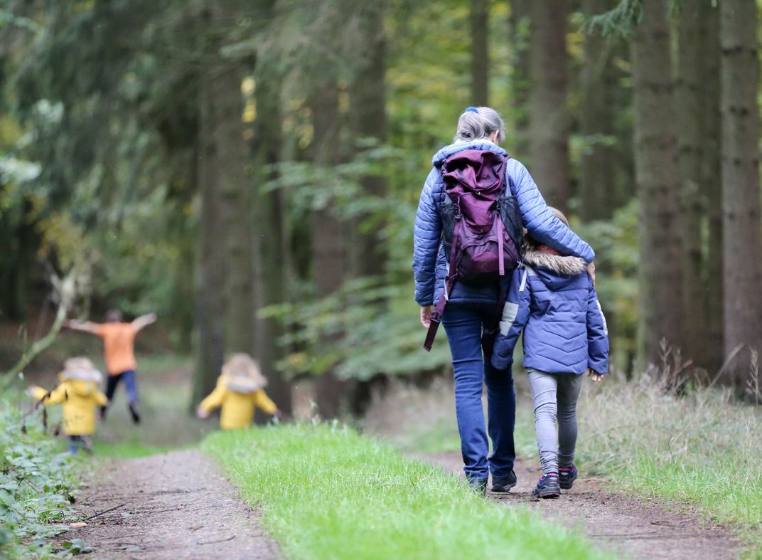 An older woman and young child in hiking clothes walk together on a forest path while three more children dash ahead.