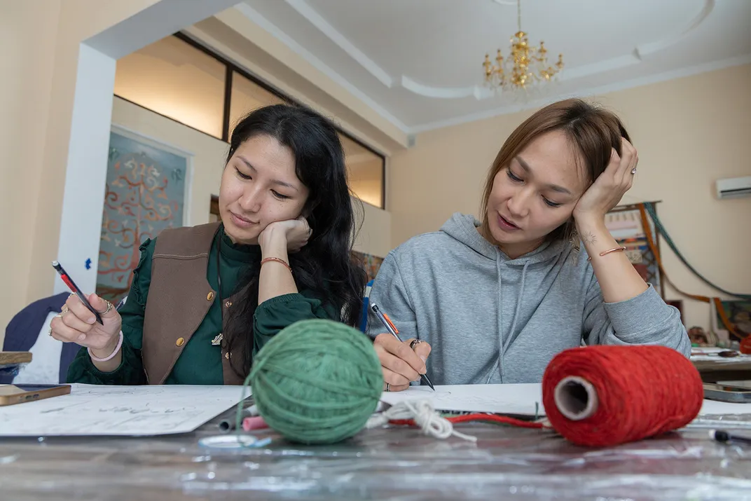 Two women sitting next to each other at a table both lean their heads on their left hands while drawing with their right. In front of them, skeins of red and green yarn.