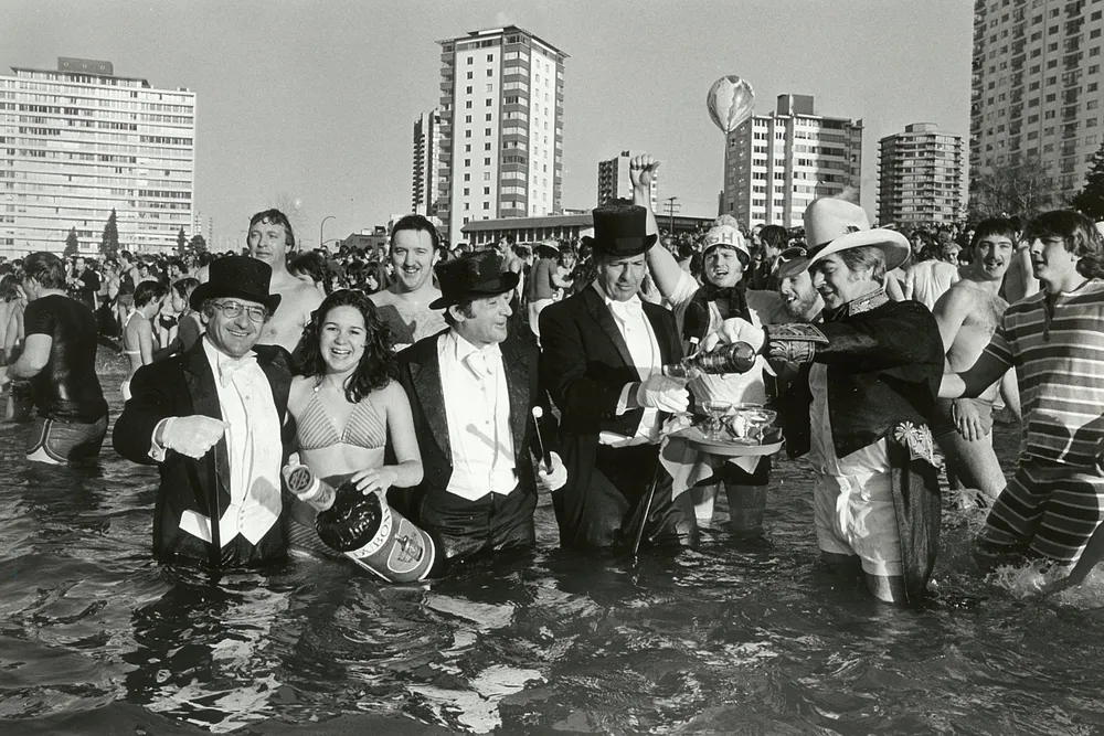 people, some in formal wear others in costumes or swimsuits, pose for a photo in water in front of a city skyline