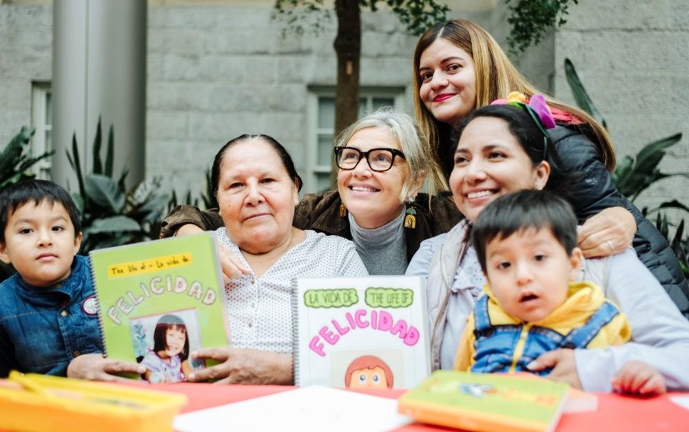 Woman smiles at the center of a group with young children and their mothers showcasing books they created