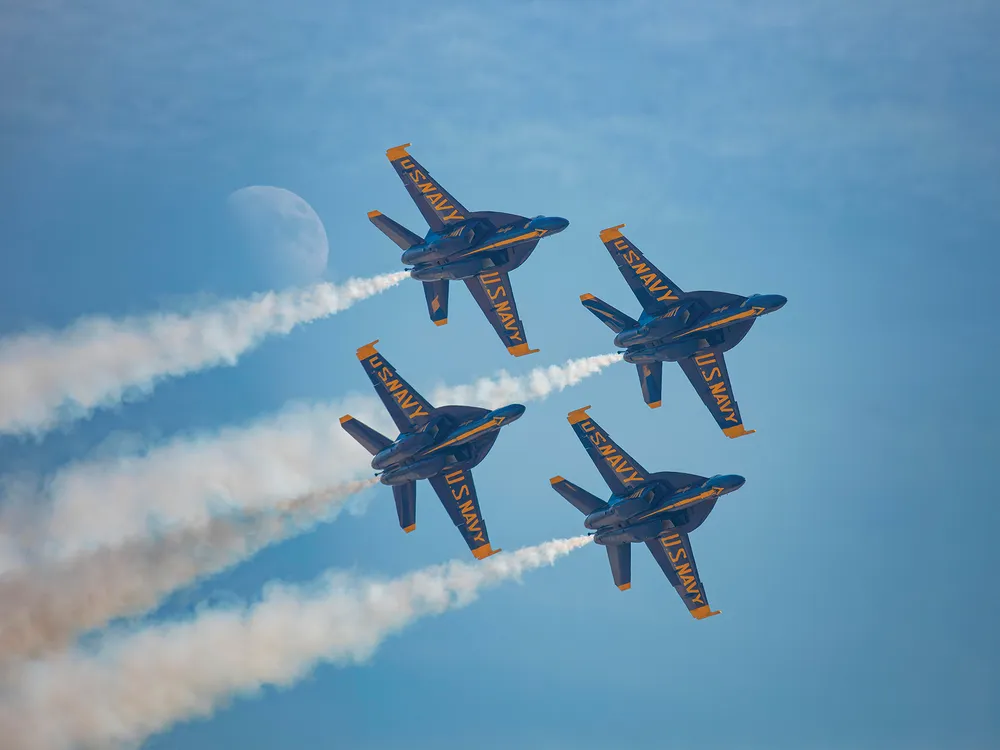 Four military jets fly in a diamond formation across a blue sky. On the underside of each jet's wings, "US Navy" is painted in bold yellow letters. White airshow smoke streams from each jet. The moon is visible in the sky.