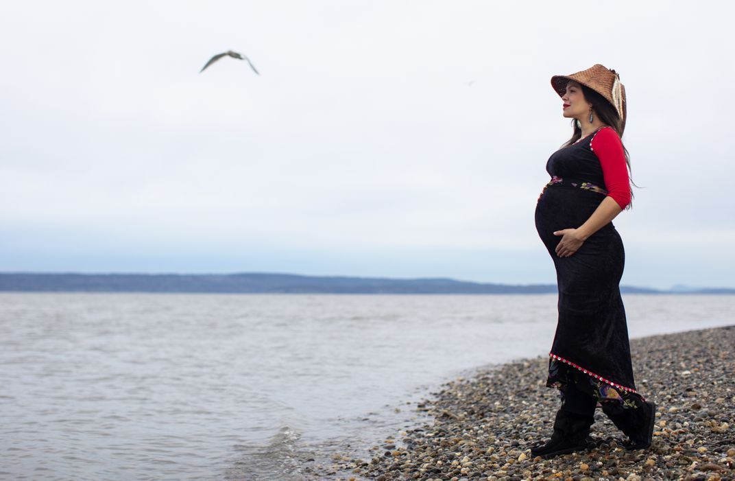 A woman in a black dress and cedar hat stands along the Salish Sea shoreline, gazing out into the distance.