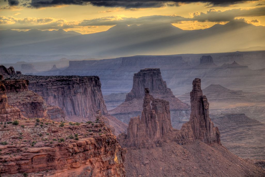 A landscape photo of Canyonlands National Park. The canyon is deep and dusty with tall, tower-like rocks jutting out of it. The sky is gold and gray, with wispy clouds.