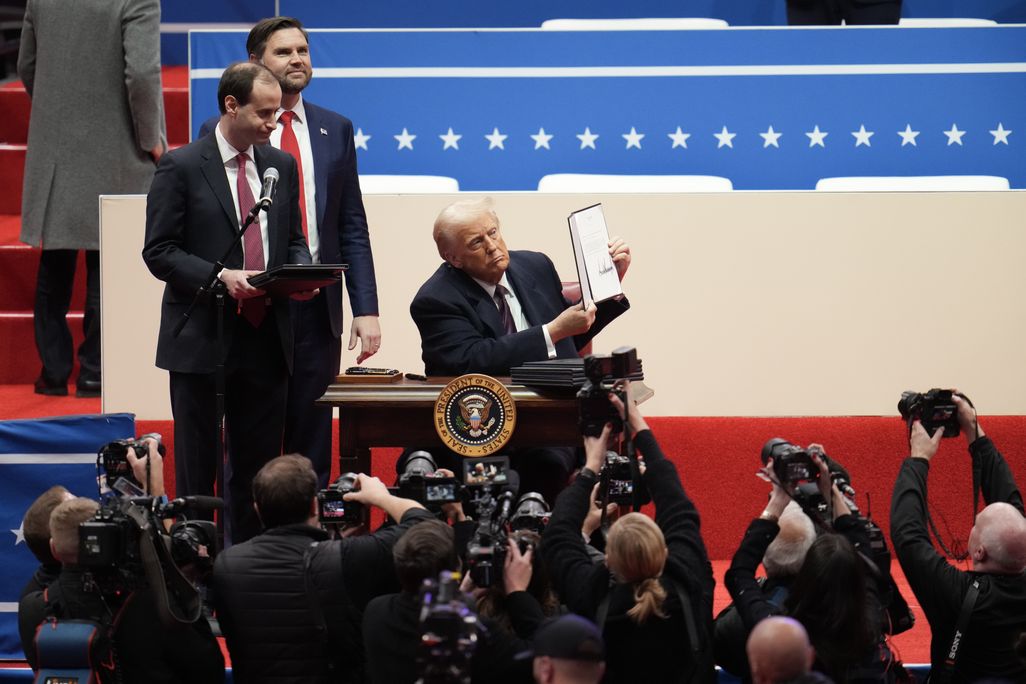 Donald Trump sits on a stage and holds up a piece of paper