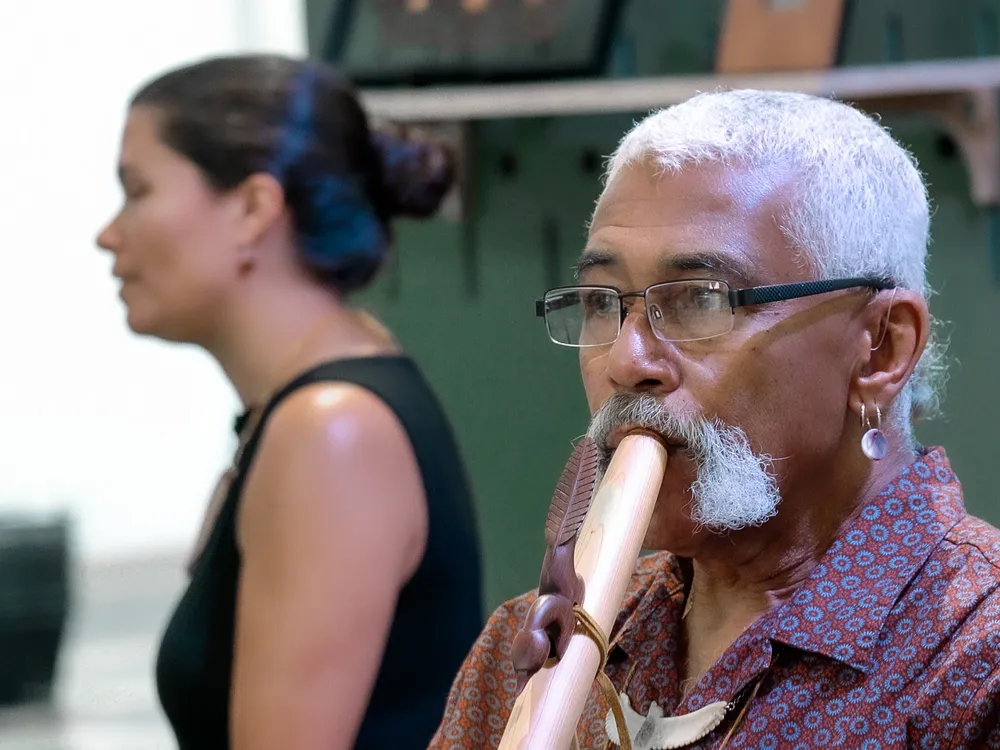 A man with short white hair, moustache, and glasses plays a wooden flute. Behind him, slightly out of focus, is a young woman with long dark hair in a bun and a black tank top.