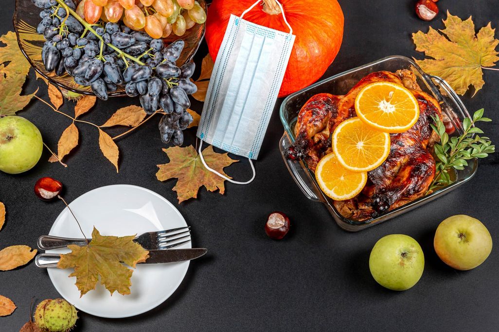 A photograph of a table taken from above. Table has dried leaves, a white plate with a fork and knife, a roasted chicken with orange slices in a glass tray. There is a a pumpkin with a blue disposable mask draped over the stem at the top of the image.