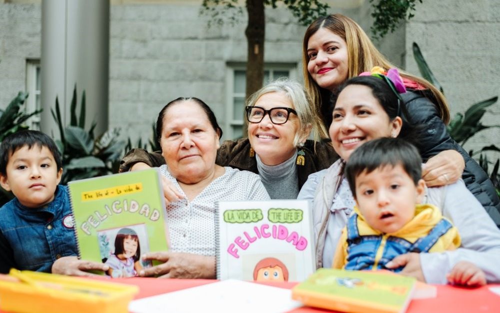 Woman smiles at the center of a group with young children and their mothers showcasing books they created