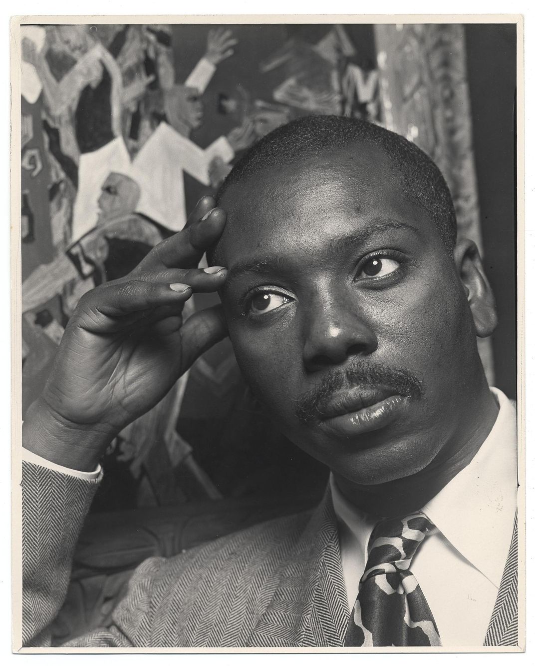A black and white image of Lawrence, a black man with a mustache, looking contemplative with a suit and tie and resting his head against his hand, in front of one of his works of art