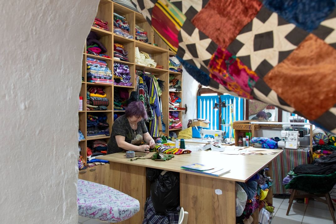 An artisan stands over a table while working on a piece of cloth. Behind her stands a shelf full of crafts and cloth. The view is partially blocked by a curved archway and a hanging curtain.