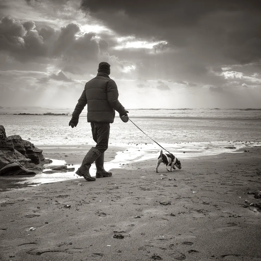 A man and dog on the beach. The dog is pulling on the lead, the sky is dark and the man is wearing Wellington boots.