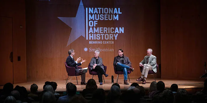 Four people appear in chairs on stage, a panel