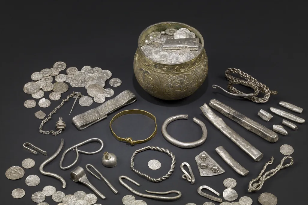 A display of silver Viking artifacts laid out on a table, including a wide mug-like cup, many coins, silver bars and what appear to be bracelets and other jewelry scattered around