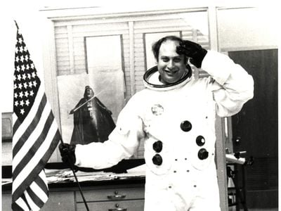 Informal classroom portrait of teacher Herbert Stephen Desind (1945-1992), wearing a reproduction Apollo-era spacesuit, holding an American flag; circa 1980s.  Desind was a space flight aficionado, and his collection of photographs of aircraft and spacecraft was donated to the National Air and Space Museum in 1997. This image is part of the Herbert Stephen Desind Collection.
