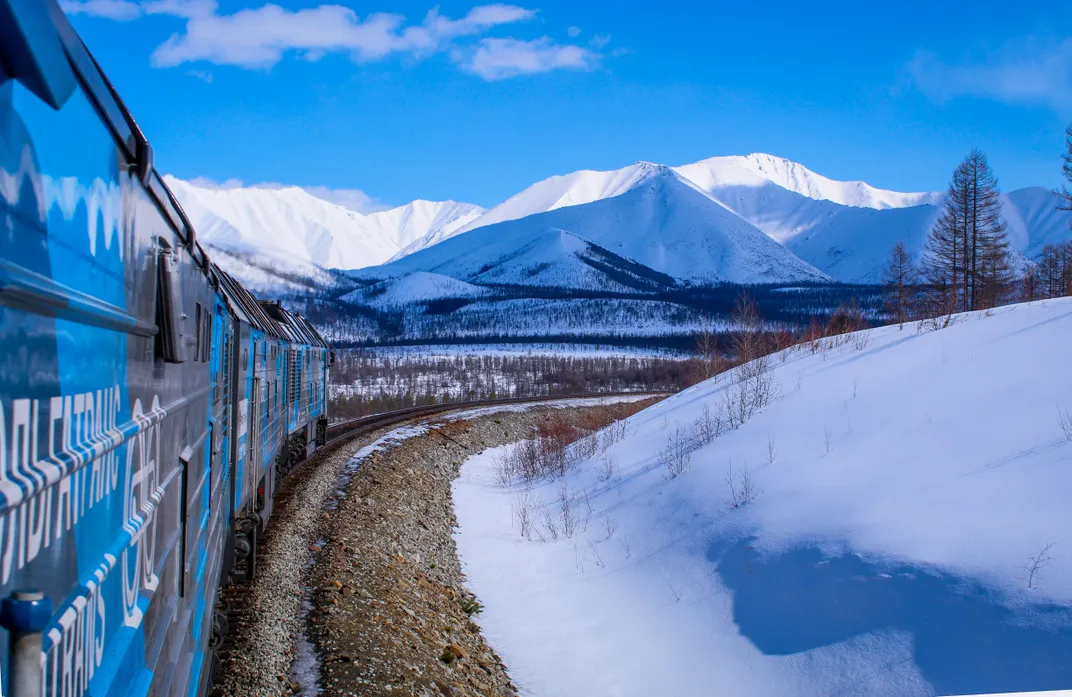 13 - A train whizzes through snowy hills with frosty peaks from the Stanovoy Ridge mountains in the distance.