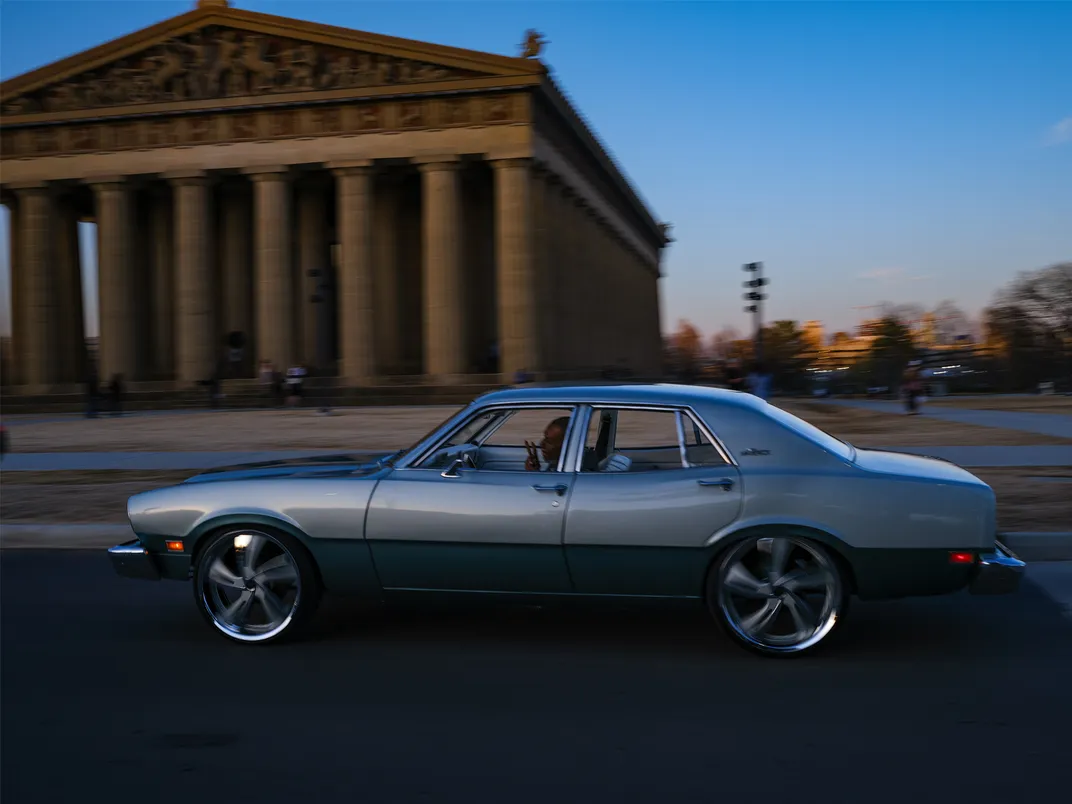 A Black man drives past the Parthenon at dusk in a blue vintage car