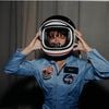 Sally Ride wearing a blue flight suit and helmet with her hands on either side of the helmet.