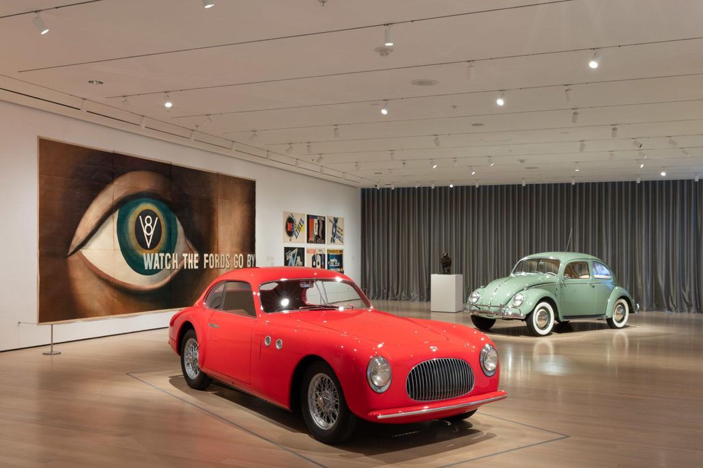 A view of a museum gallery with a bright red car on display in front of a light green Beetle; on the wall, an enormous lithograph of a human eye with the words Watch the Fords Go By