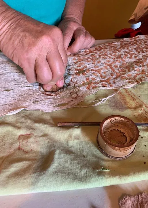 Close-up on two hands using a stamp to print a reddish brown pattern on a piece of pink bark cloth.