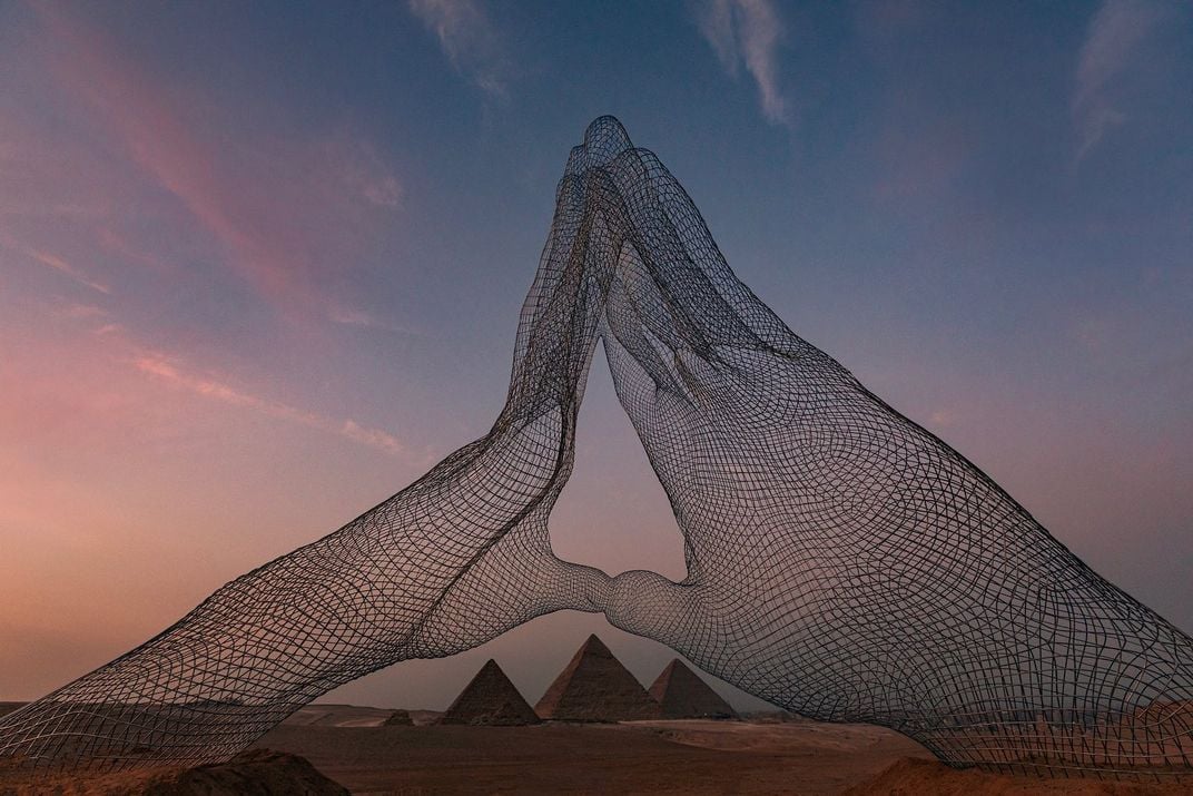 Two mesh wire structures of enormous hands touch fingertips, framed by the pyramids at dusk with a sunset behind
