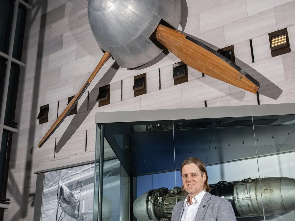 A white man in his late 30s stands inside a gallery displaying aviation artifacts, including a four-blade fan mounted on the wall.