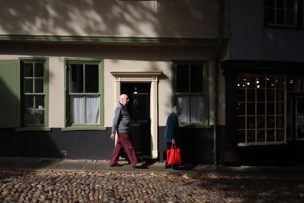 An elderly couple walks through a quiet, cobblestoned street in a historic English town, illuminated by patches of warm autumn light. The traditional architecture, painted windows, and soft shadows create a timeless atmosphere that reflects the charm of exploring small European towns on foot. This moment captures the peaceful rhythm of local life that travelers often discover beyond the busy landmarks.