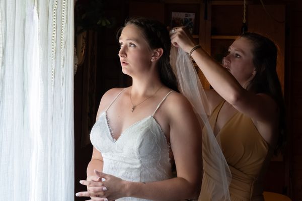 A bride stands near a window as her sister puts on her veil. thumbnail