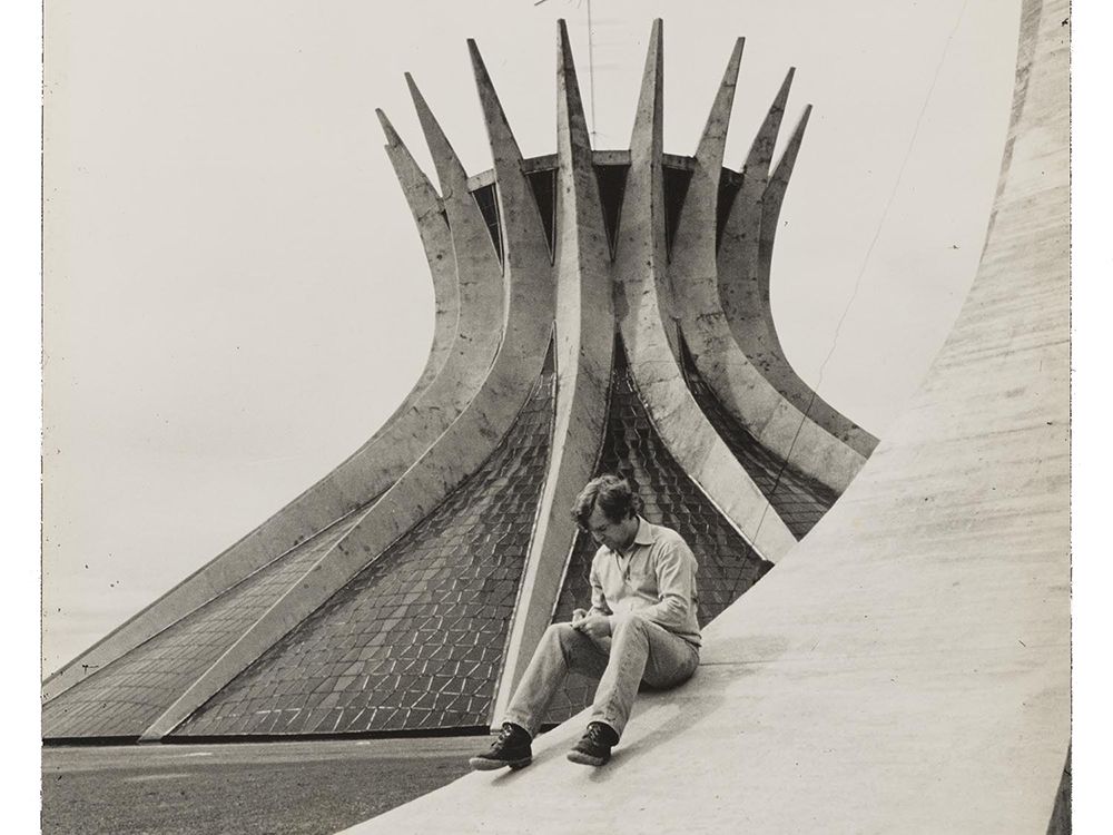 Grayscale image of Robert Hughes sitting on the sloping curve of a building examining something in his hands, with a modernist church in the background.
