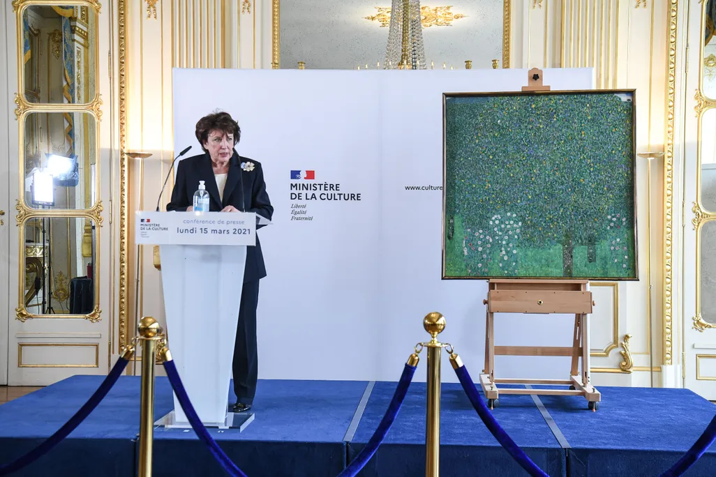 woman with brown hair and blue pantsuit standing at podium in front of pastel floral painting