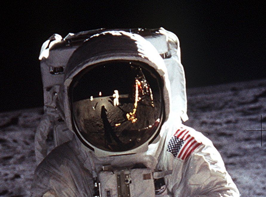 Astronaut Buzz Aldrin wears a large helmet and white space suit while standing on the moon. The American flag, Neil Armstrong and more of the moon is reflected in his helmet. The moon's rocky, gray surface makes up the background.