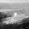 An old photo, tinted brown, depicts a flat barren landscape with a single pillar of dense white smoke at the center of the photo.