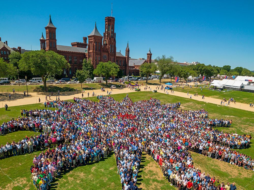 Thousands of people assemble on the green grass of the National Mall to form a sunburst shape, the Smithsonian logo. The red-brick Smithsonian Castle rises in the background.