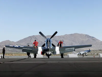 Speedball Alice, a restored dark green P-51D airplane with white stripes, is parked on a tarmac in the Reno desert, with mountains and blue sky in the background.