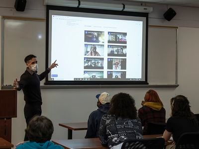 A man points to a video screen in a classroom with students seated in the foreground.