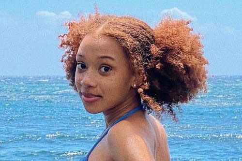 Portrait from shoulders up of a girl in front of the ocean smiling. Her hair is a reddish-orange hue, curly, and naturally styled into pigtails.
