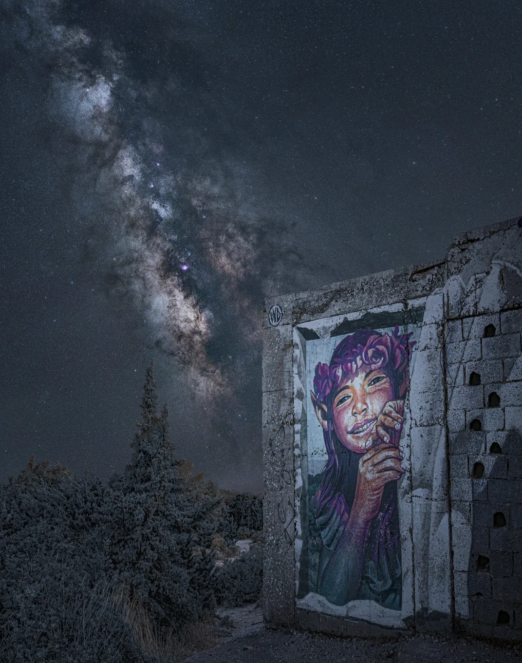 the milky way stretches across the sky, with a painting of a young woman with a purple crown of flowers in the foreground on a wall of a ruin