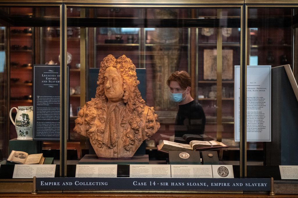 A glass case with the newly positioned bust of Sloane, which is a terracotta color, next to a small sign that has paragraphs of text explaining how he profited from enslavement. Behind, a visitor walks by wearing a mask.