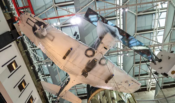 A World War II military airplane is photographed from below while hanging from the ceiling. The underside of the airplane is painted white and the wing and forward fuselage are wrapped in opaque plastic.