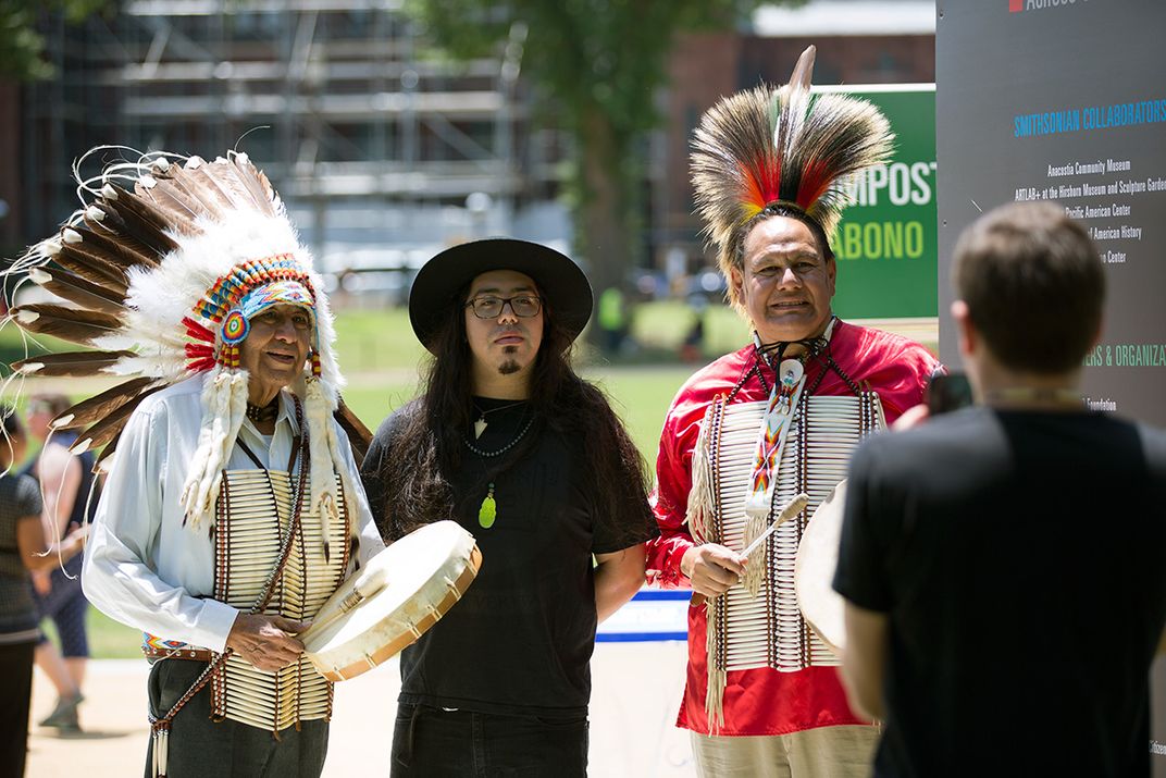 Three men stand together, smiling. The men on the left and right are wearing traditional feathered headdresses and the man in the middle has long hair and wears a black, wide brimmed hat.