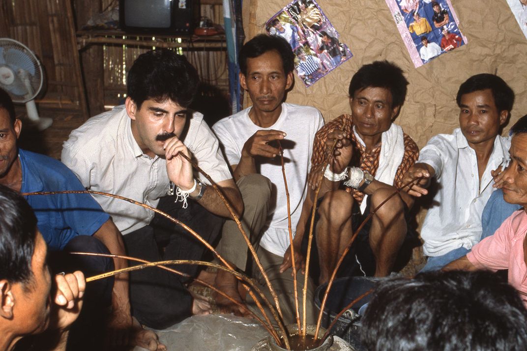 A group of men drink from a central beverage cup using long, bending shoots of bamboo like straws.